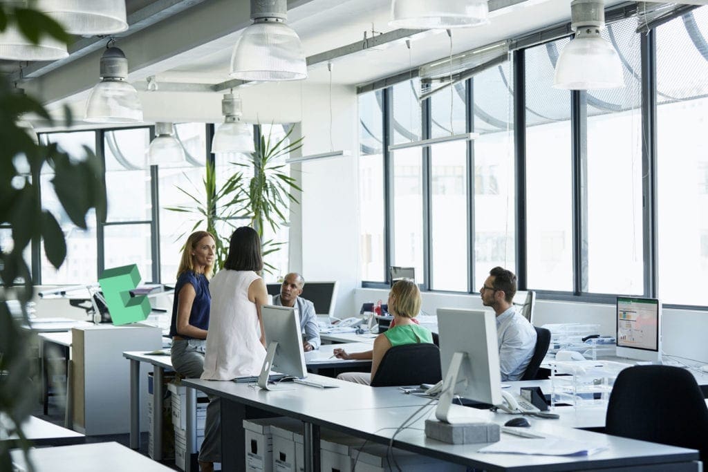 Group of men and women sit around a table in open-plan office, talking business-critical